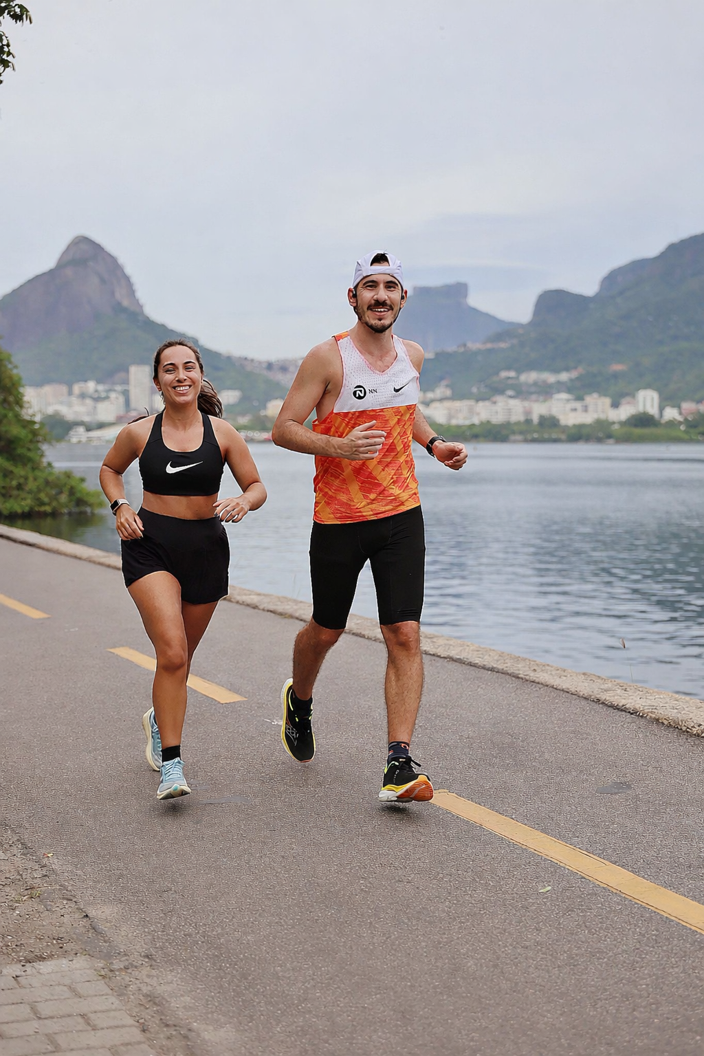 Casal de corredores de rua ao lado de um lago, com montanhas ao fundo, durante o dia.