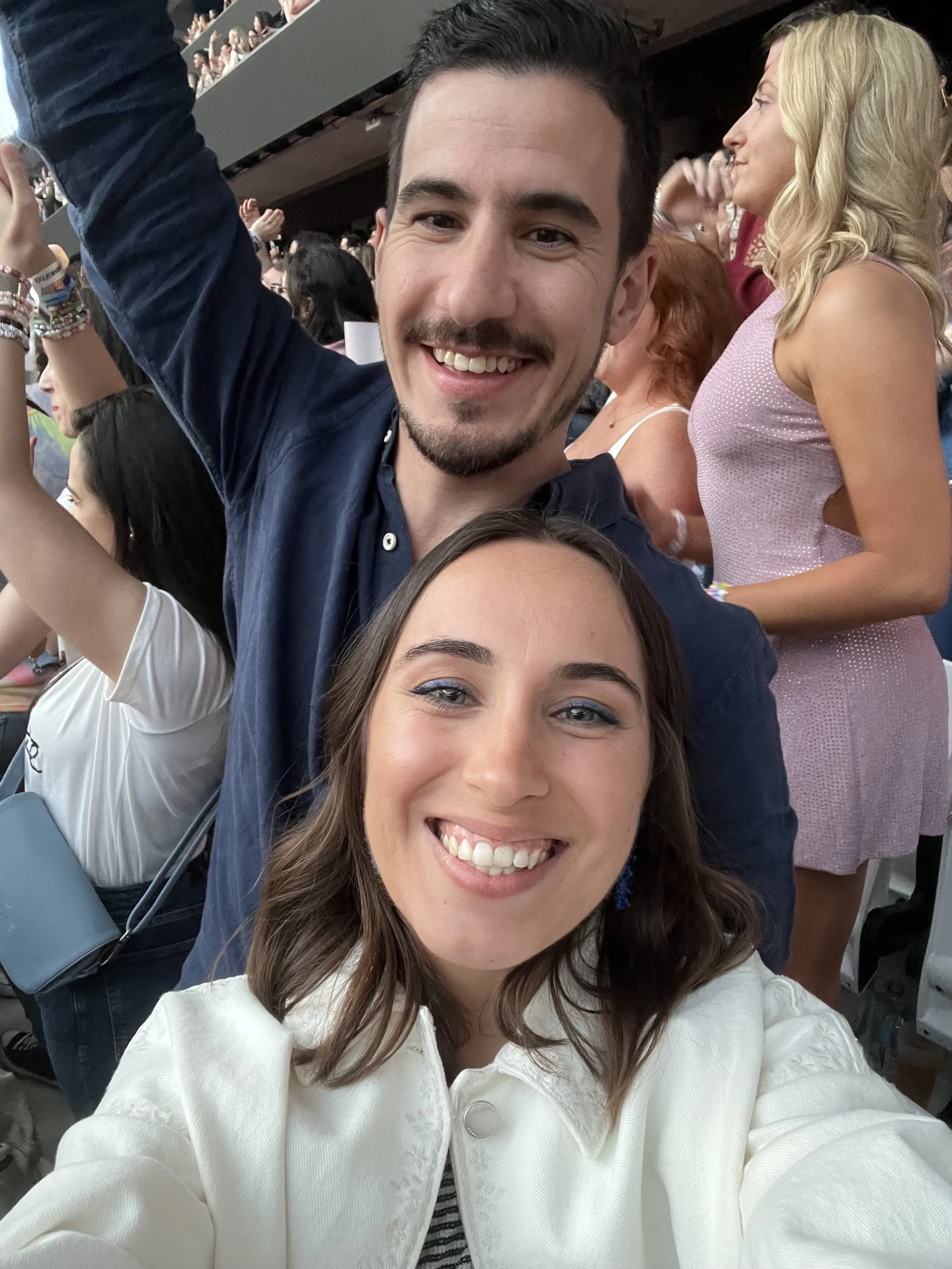 Homem e mulher sorrindo enquanto tiram selfie em evento com público ao fundo.