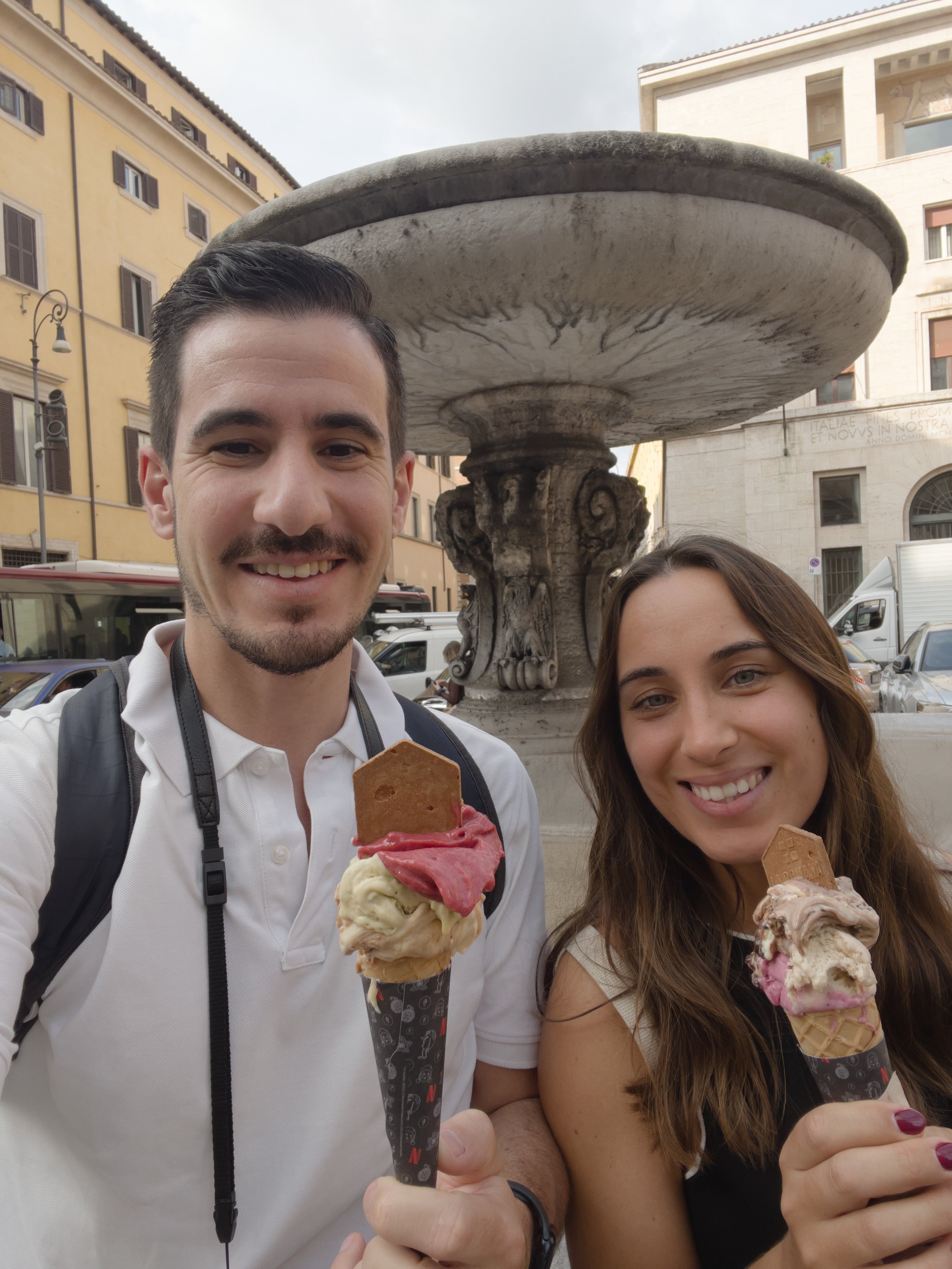 Homens e mulher sorrindo segurando casquinhas de sorvete em frente a uma fonte em uma praça urbana.