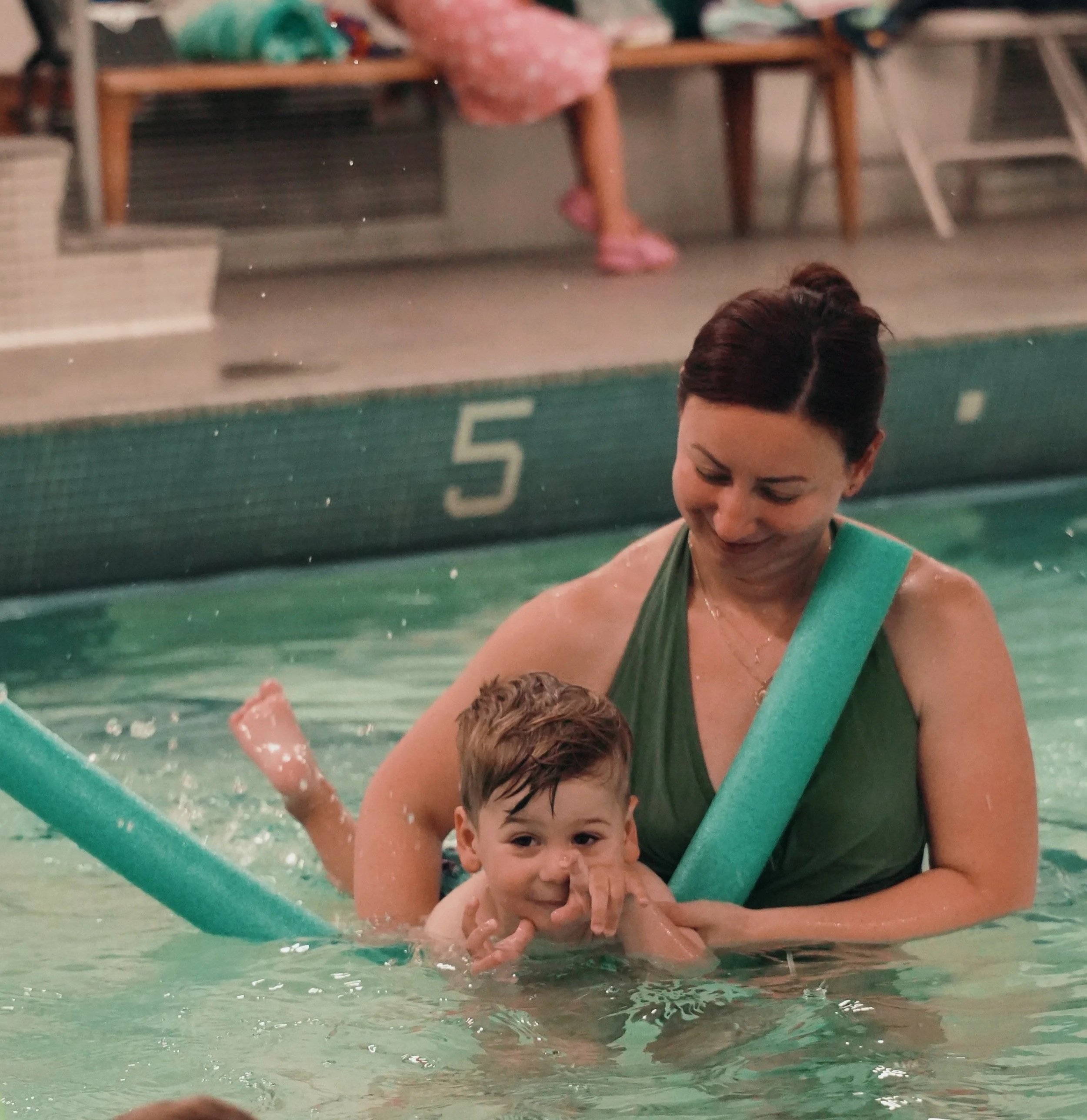 A woman and a young boy, likely a mother and son, enjoy swimming together in a pool. The woman is smiling and holding the boy, who is in the water and looking at the camera with one hand near his face.