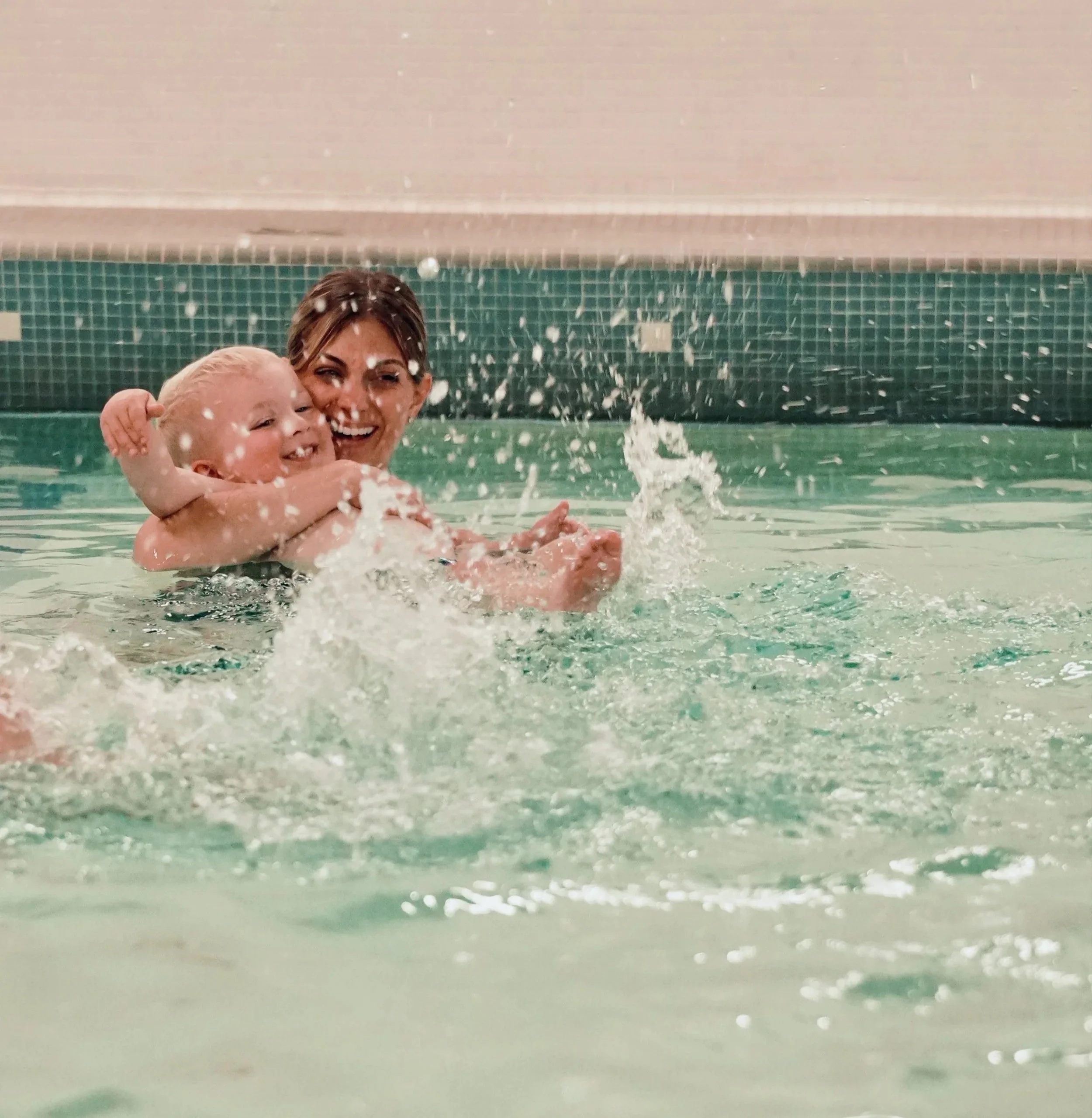 A woman and a young child swimming and playing together in an indoor pool, smiling and splashing water.