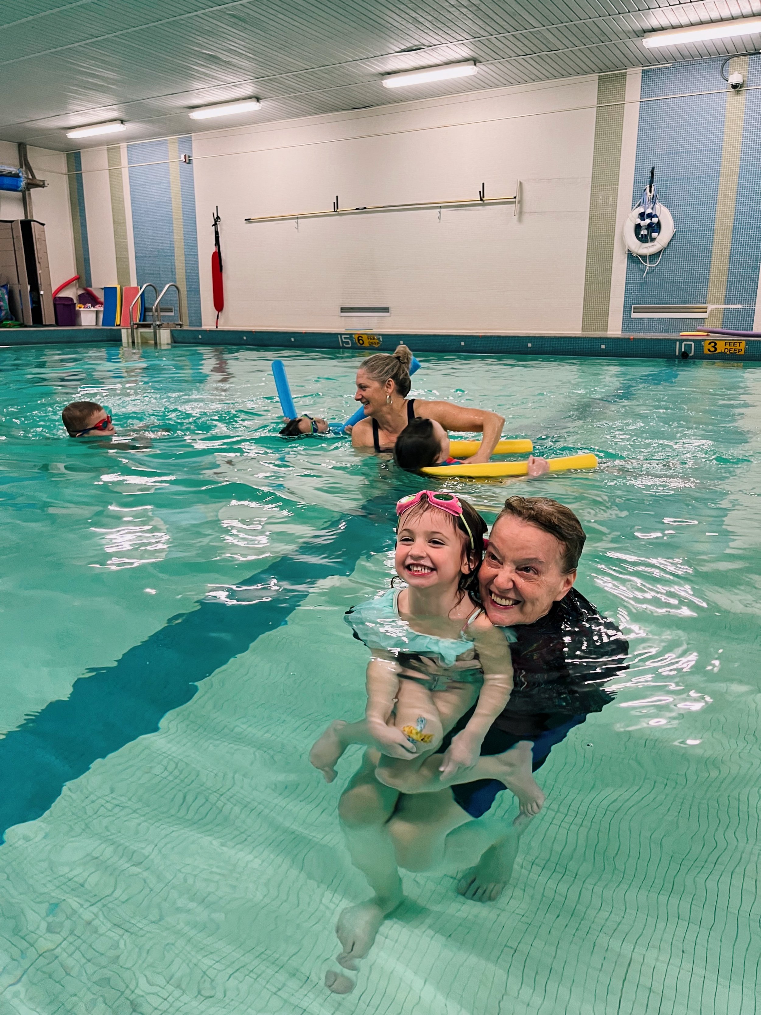 A happy group of children and adults enjoying a swimming lesson in an indoor pool, with some using floatation devices and swimming gear.
