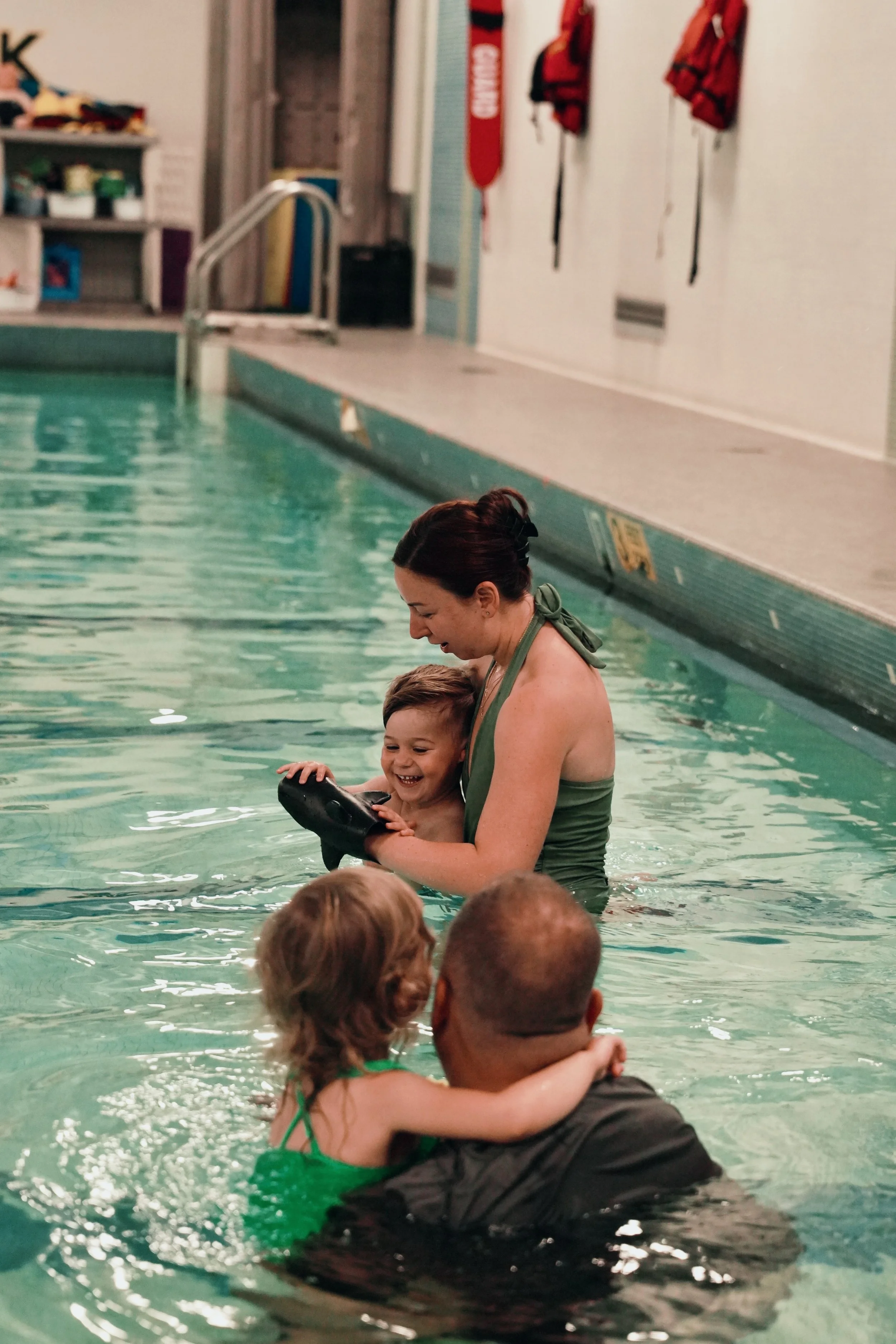 A woman teaching children how to swim in an indoor pool, with a young boy smiling and a girl hugging an adult man.