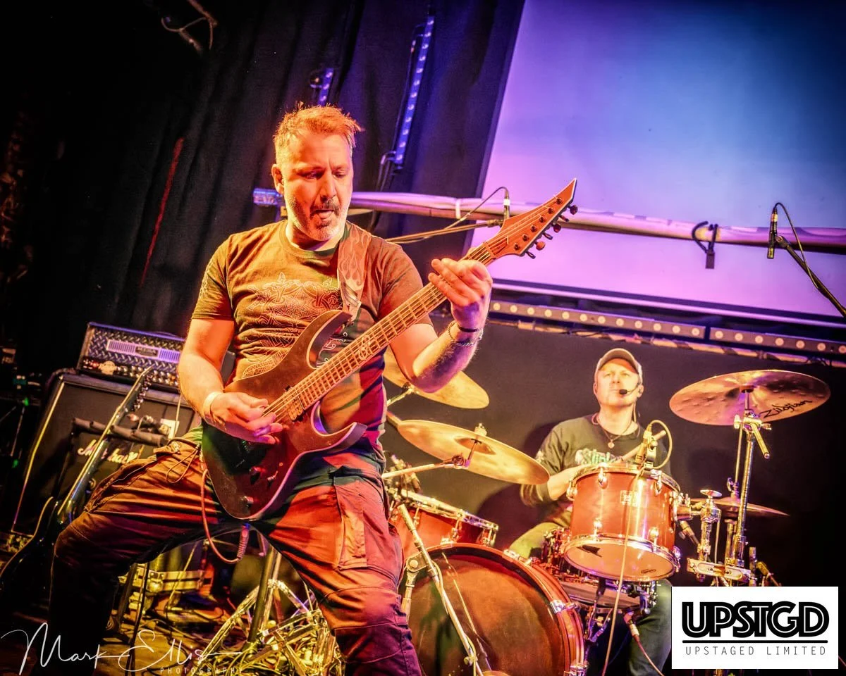 Two musicians performing on stage; one playing an electric guitar and the other on drums during a concert.