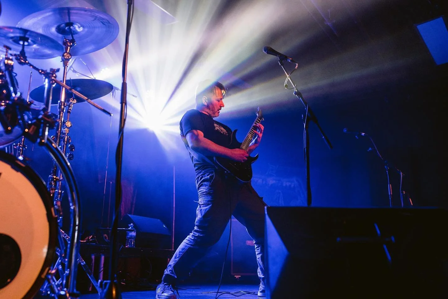Musician playing electric guitar on stage with colorful lighting and drums visible on the side.