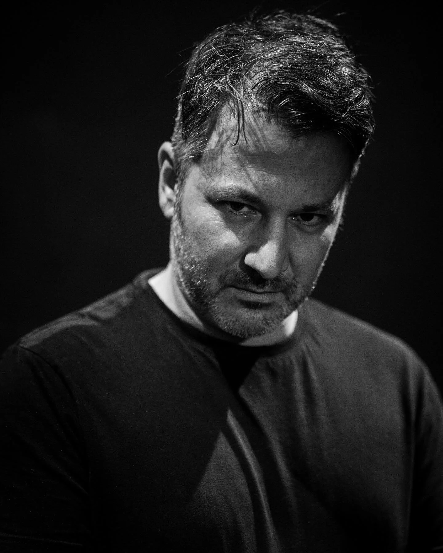 Black and white close-up portrait of a man with styled hair and a beard, looking down with a serious expression against a dark background.