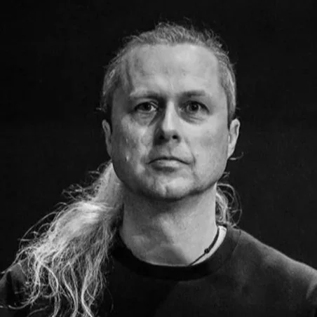 Black and white photo of a man with long hair tied back, wearing a dark shirt, looking directly at the camera.