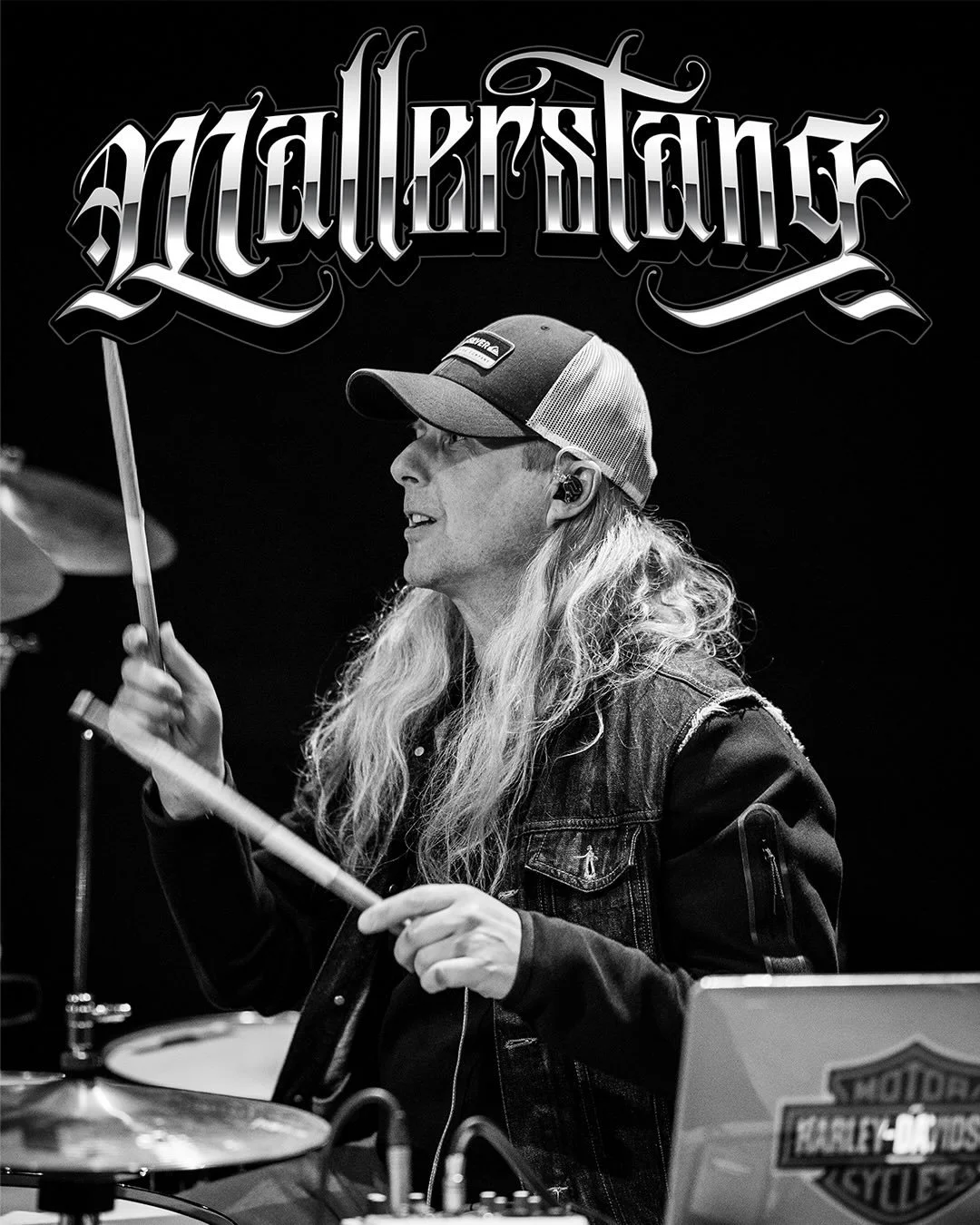 Black and white image of a male musician with long hair wearing a cap, playing drums at a Harley-Davidson event.