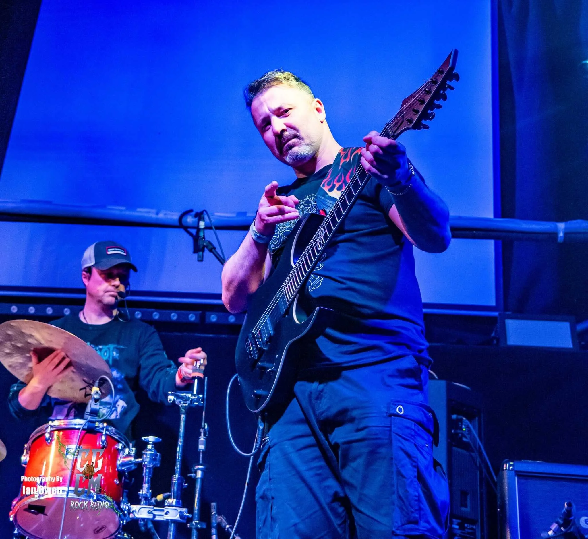 A man with short hair and a beard is playing an electric guitar on stage. He is pointing at the camera and making a finger gun gesture, wearing a black T-shirt with flame designs on the shoulders. In the background, another musician is playing drums, wearing a baseball cap and headphones, in a dimly lit, blue-toned concert venue.