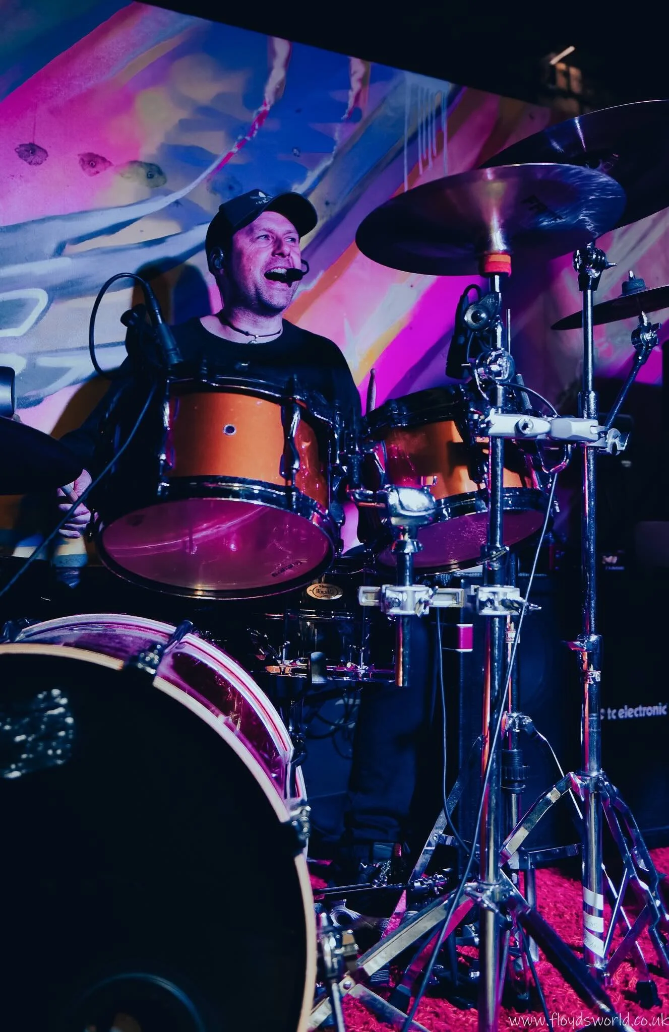 A man playing electronic drums at a concert or performance, with colorful lighting and a vibrant background.
