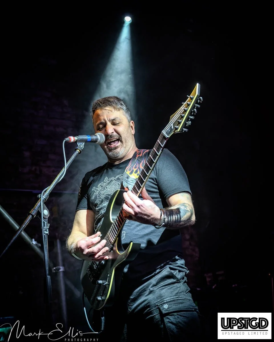 Man playing electric guitar and singing into a microphone on stage with a spotlight overhead.