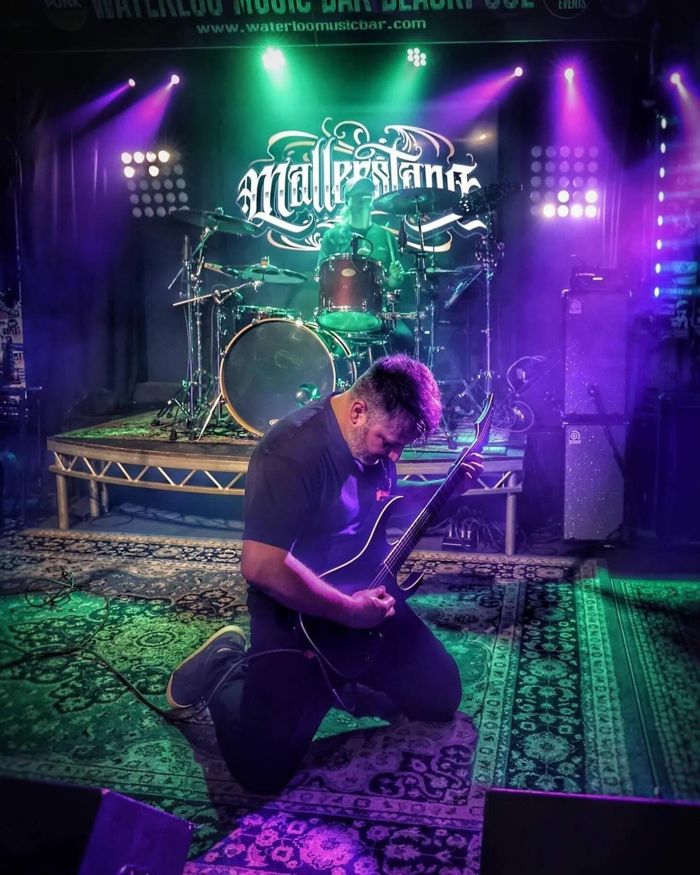 A musician kneeling on stage playing an electric guitar with a drummer in the background, illuminated by colorful stage lights. The stage has a dark backdrop with the word 'MetalPasta' written on it and a website URL at the top.