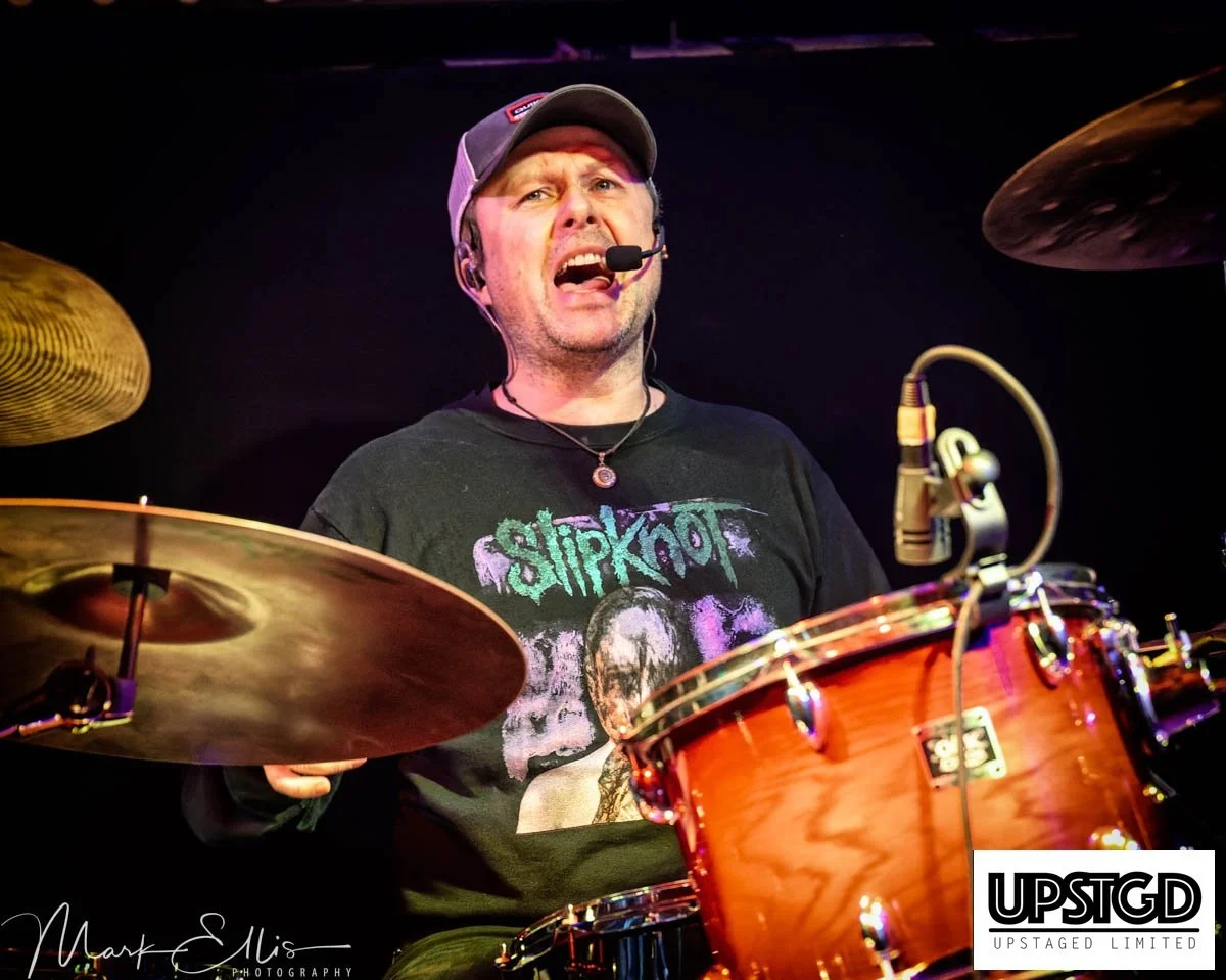 A man playing a drum kit on stage, singing into a headset microphone, wearing a Slipknot t-shirt and a gray baseball cap.