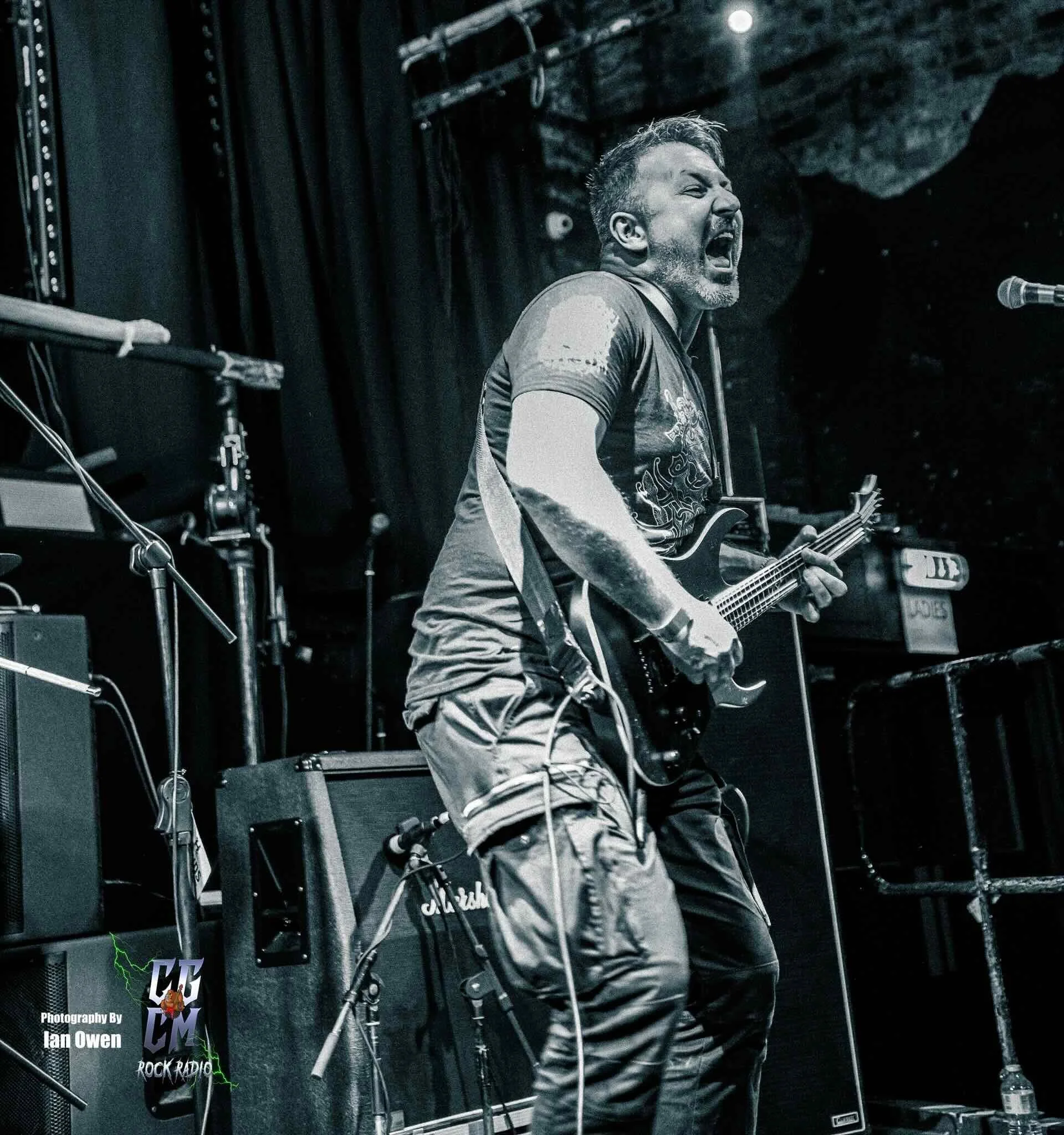 A guitarist passionately performs on stage, singing into a microphone with his eyes closed, dressed in a dark t-shirt and cargo pants, surrounded by sound equipment and stage lights in a black and white photograph.