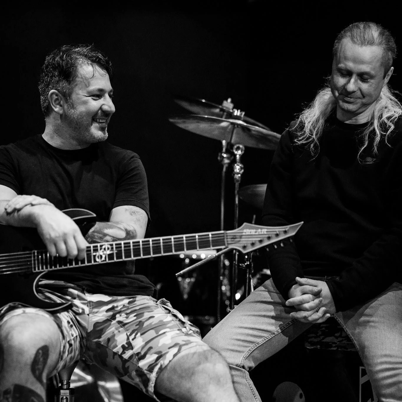 Two men sitting and talking, one holding an electric guitar, with drums in the background.