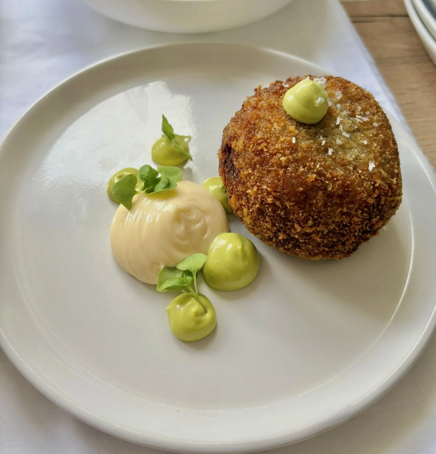 Fried croquette with green sauce on top, served with a dollop of creamy white sauce and garnished with microgreens and small green vegetables on a white plate.