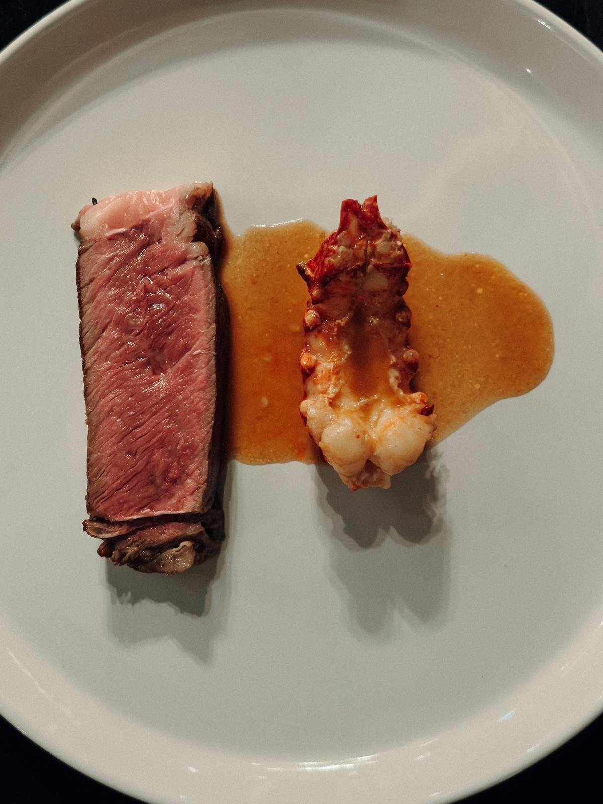 A plate with a piece of medium-rare steak, a lobster tail with sauce, and a pool of sauce beneath it.