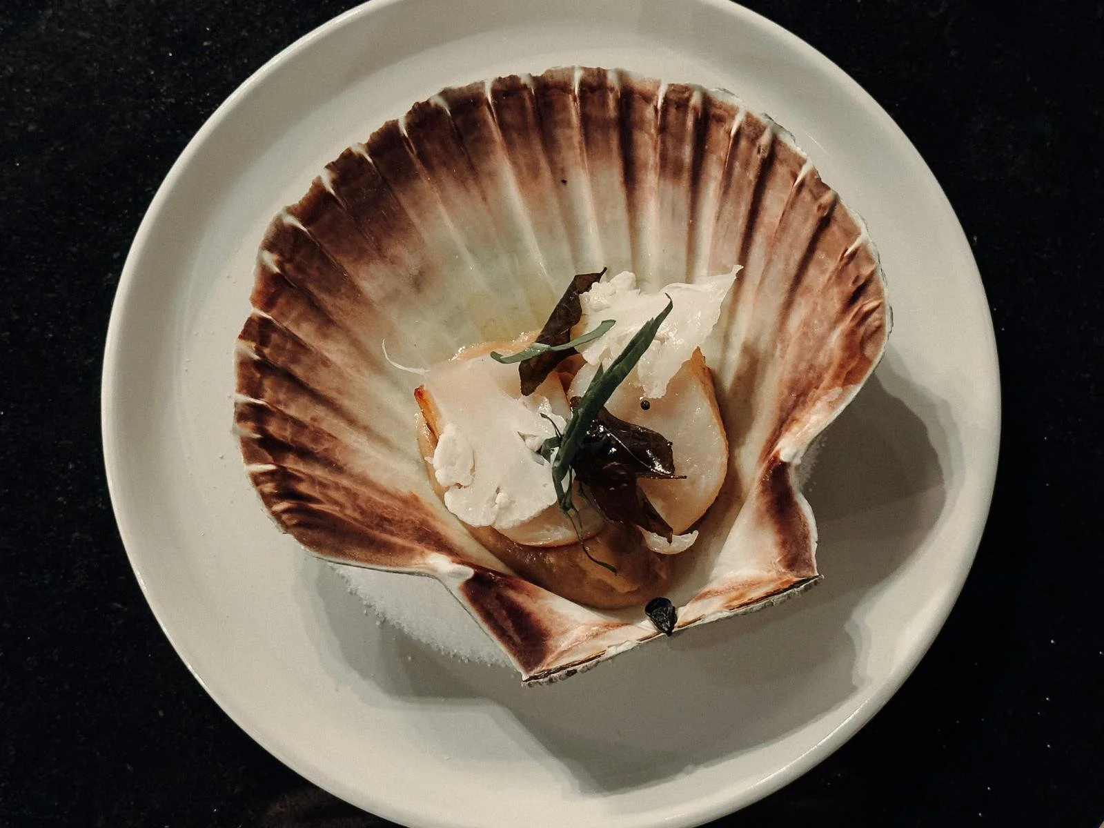 A scallop shell filled with a cooked scallop topped with herbs and garnishes, served on a white plate.