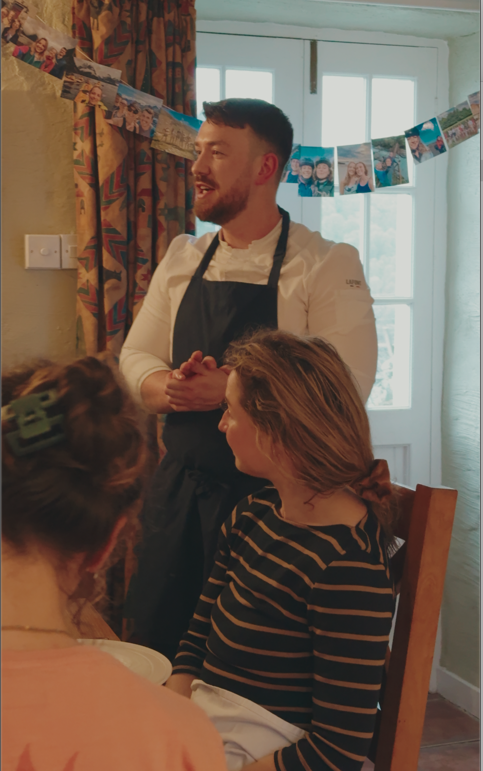 A man wearing a white shirt and black apron speaks to two women sitting at a table in a cozy, decorated room. One woman has shoulder-length hair and wears a striped shirt, while the other has her hair in a bun with a large green hair clip. In the background, a string of photographs hangs across the room near a window, which has a white frame and patterned curtains.