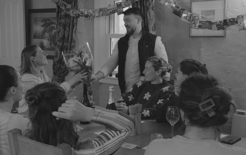 A man stands smiling at a birthday celebration with women seated around a table, some reaching for a gift.