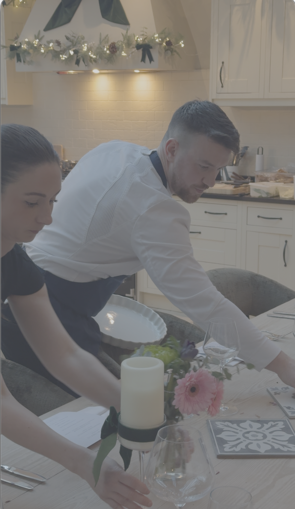 Two people setting a dinner table in a kitchen, with candles and flowers as decoration.