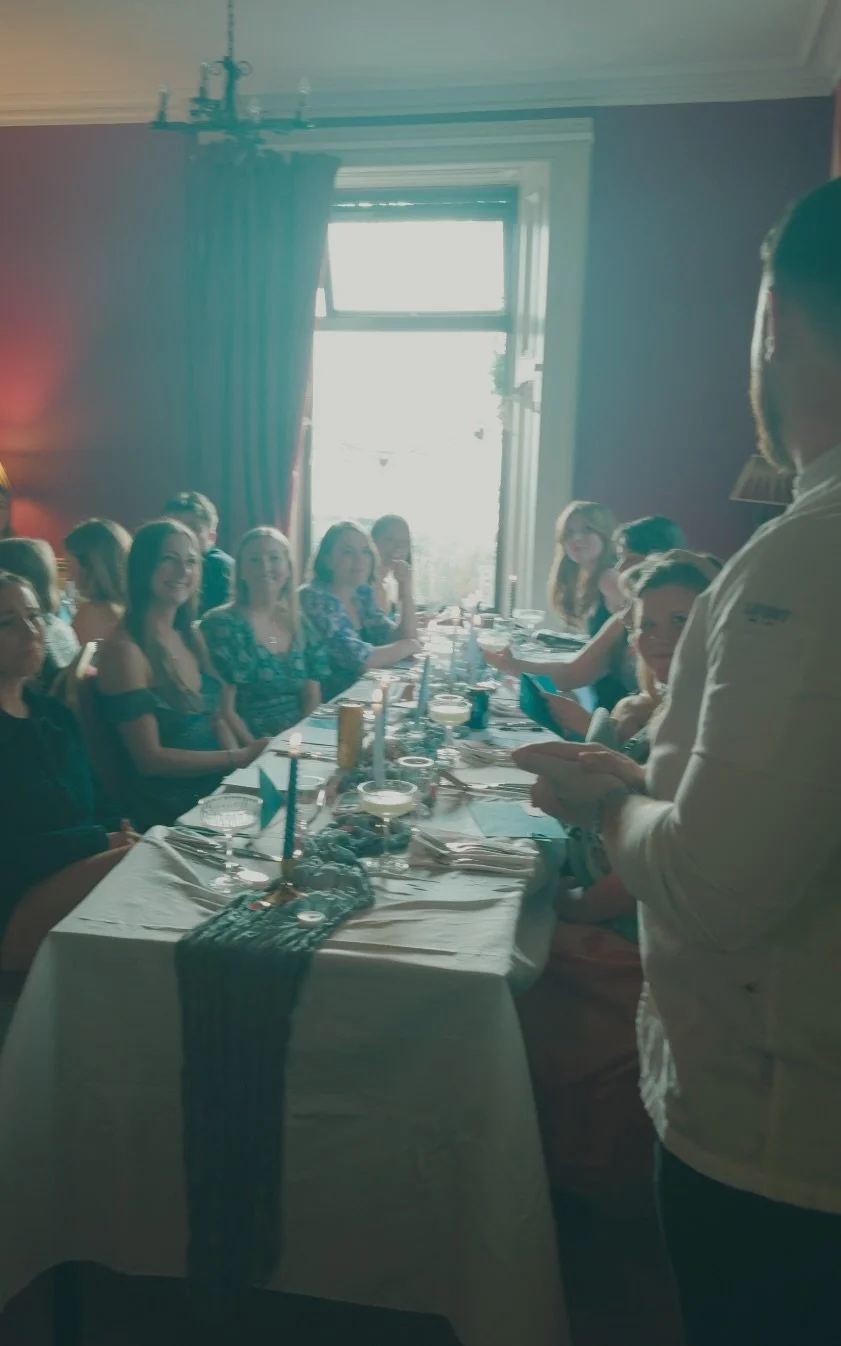 People sitting at a long dining table during a meal in a well-lit room with large windows and blue curtains, with a man standing and speaking to the seated guests.