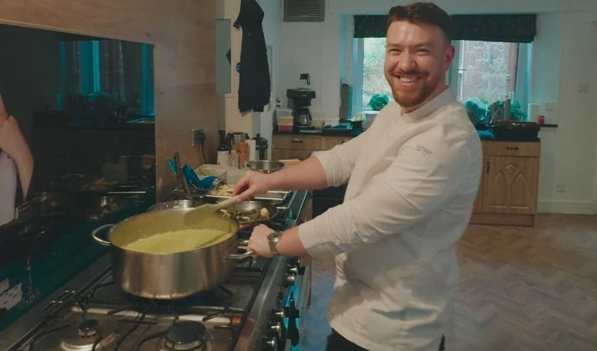 A man with a beard and a smile cooking pasta in a pot on a stovetop in a kitchen.