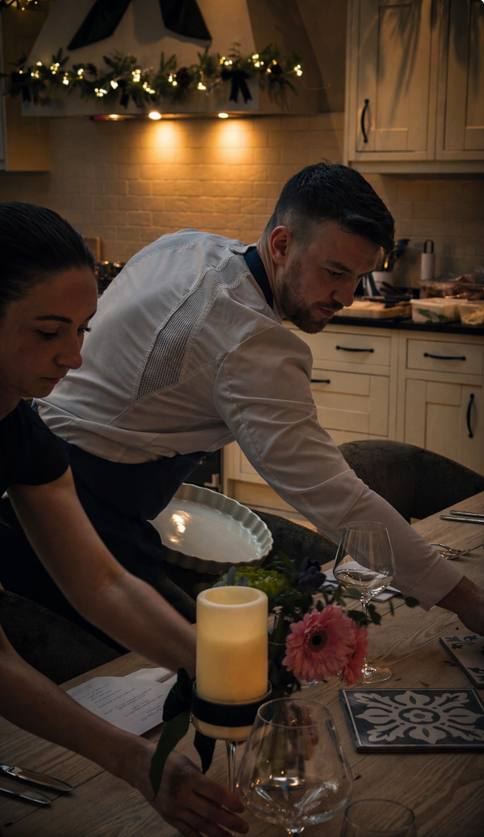 People setting a table in a cozy, dimly lit kitchen decorated for the holidays.