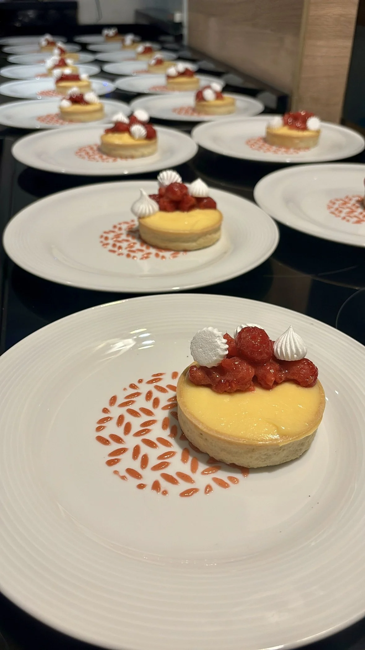 Multiple white plates each with a small round cheesecake topped with strawberry compote, dollops of whipped cream, and decorative red sugar design, arranged on a black countertop.