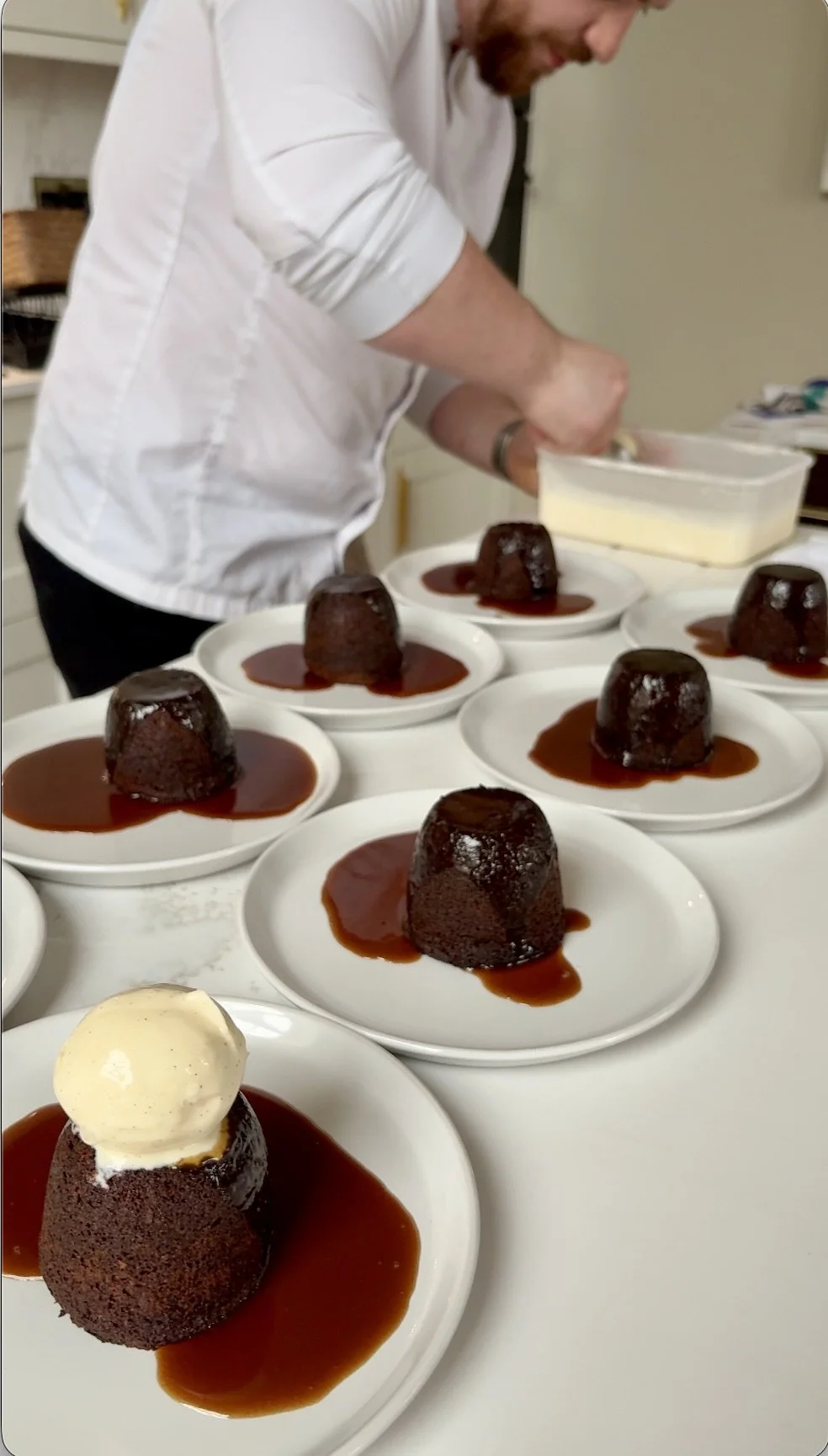 A person in a white shirt preparing multiple chocolate lava cakes topped with vanilla ice cream and drizzled with caramel sauce on white plates.
