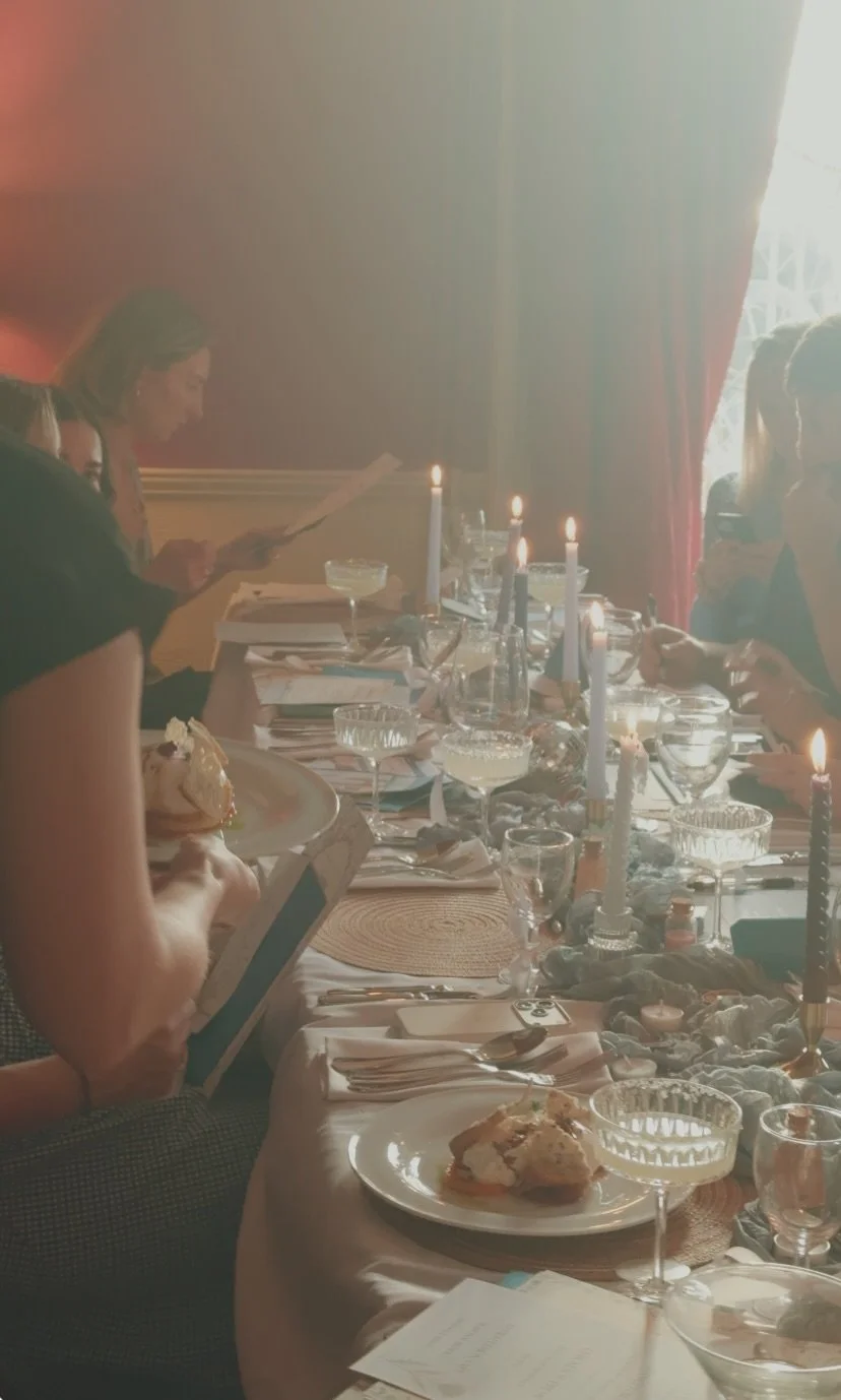 A table set for a meal with plates of food, glasses, candles, and people in a dimly lit room with red curtains.