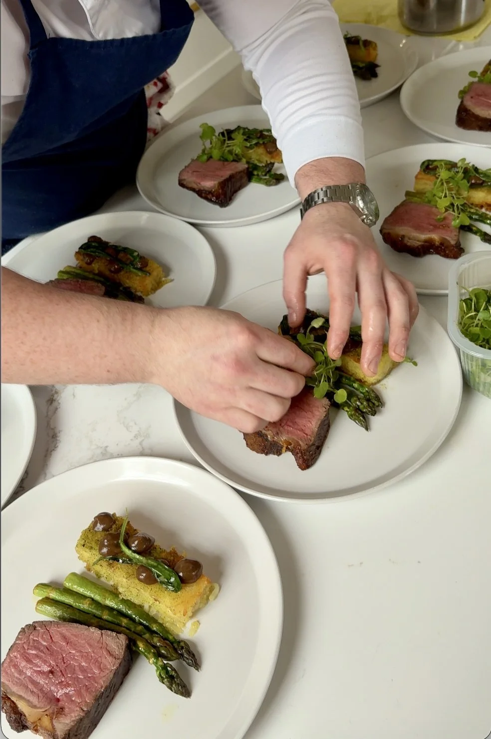 People preparing plates with grilled steak, roasted asparagus, and garnished with greens in a professional kitchen.