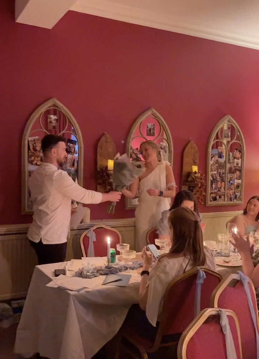A woman in a white dress is receiving a bouquet of flowers from a man in a white shirt during a celebration in a decorated dining room with pink walls and framed photographs.