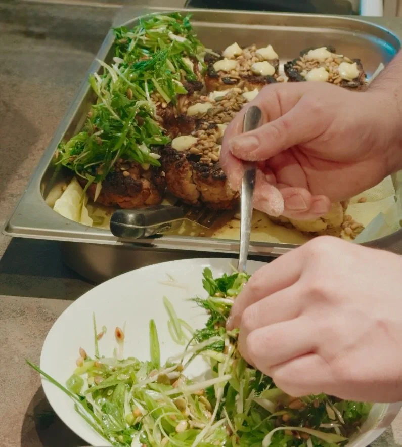 Person garnishing a plate of salad with fresh herbs, with a tray of roasted chicken topped with nuts and greens in the background.