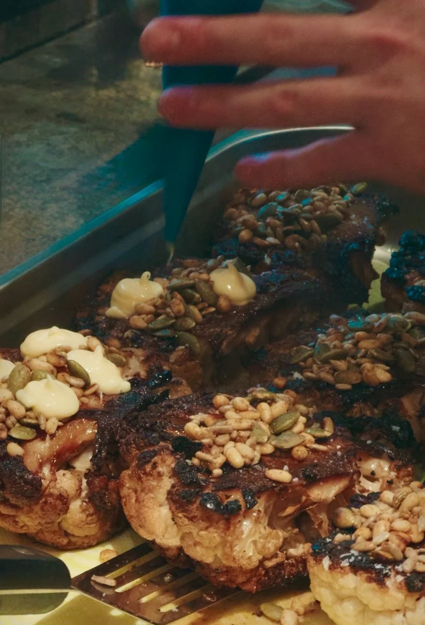 A person slicing grilled cauliflower topped with chopped nuts and a creamy sauce, on a plate.