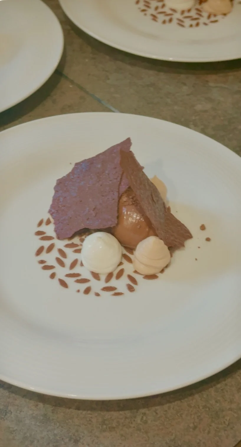 A dessert featuring a slice of chocolate cake with a thin layer of chocolate icing, garnished with two dollops of whipped cream or mousse, on a decorative white plate with a brown leaf pattern.