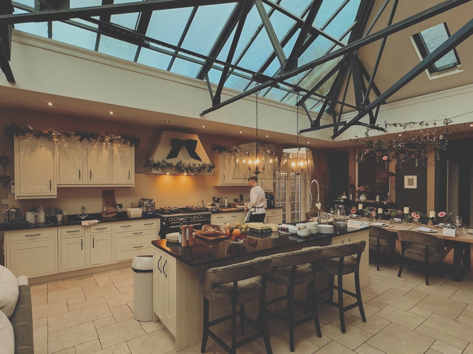 A spacious kitchen with cream-colored cabinets, black countertops, and a large island. A chef is preparing food. The room has a glass ceiling and a decorated fireplace, with dining tables set with flowers and candles.