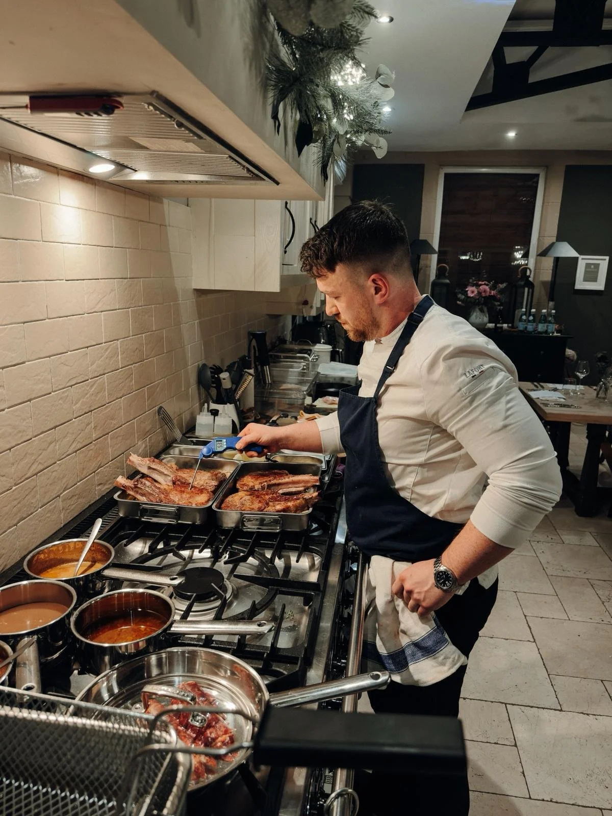A chef in a white shirt and dark apron is cooking meat at a stove in a restaurant kitchen.There are pots and kitchen tools around him and a decorated counter above.