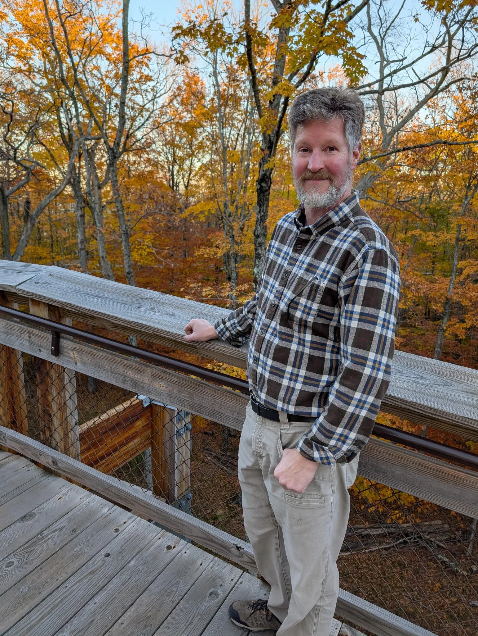 A man with gray hair and a beard stands on a wooden observation deck, holding the railing, with a background of fall foliage in orange, yellow, and brown in a forested area.