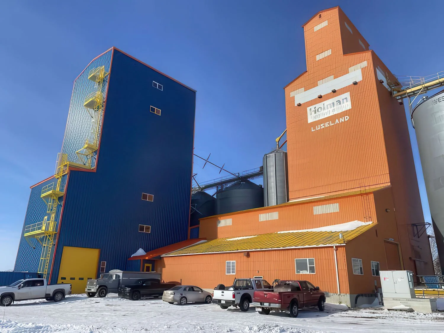 Colorful industrial building with blue, orange, and yellow sections, and several parked cars in a snow-covered lot under a clear blue sky.