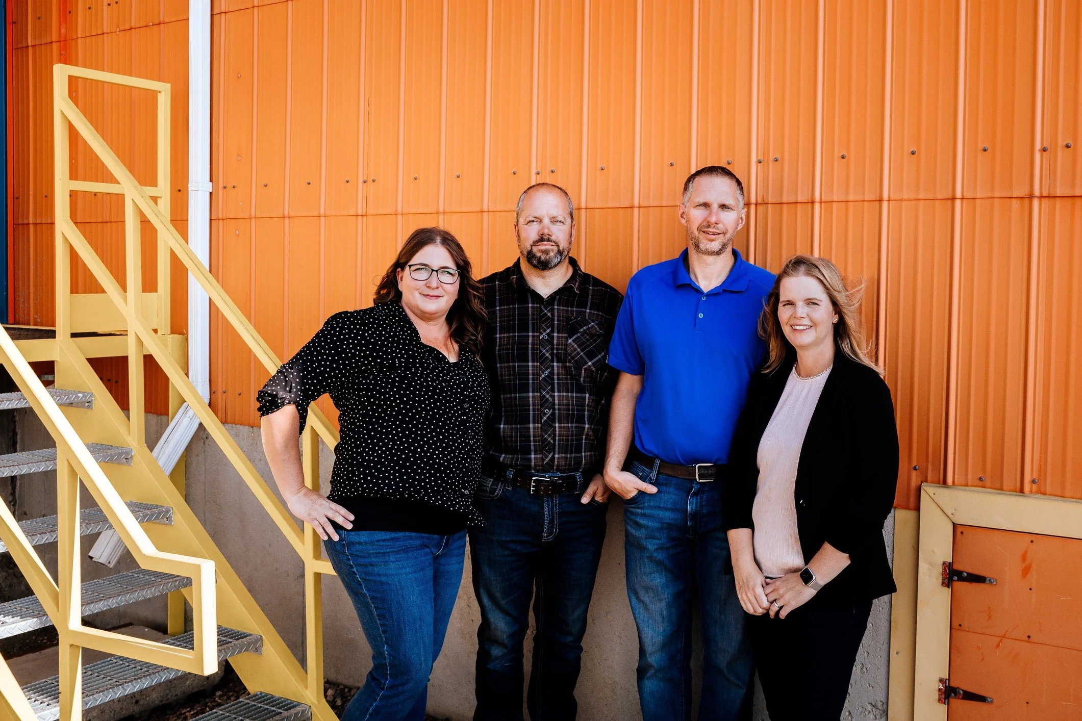 Group of four adults standing outdoors in front of an orange building with yellow stairs. The group includes two women and two men, all smiling and facing the camera.