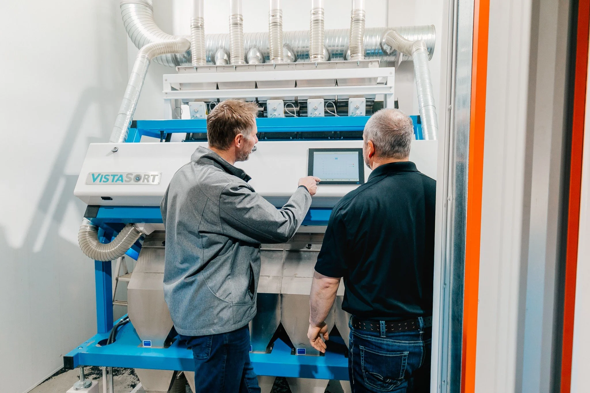 Two men, one in a gray jacket and the other in a black shirt, operate a machine labeled Vista SORT in a modern industrial setting. One man is pointing at a computer screen attached to the machine.