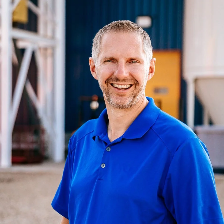 A man with short gray hair and a beard smiling, wearing a blue polo shirt, standing outdoors with a blurred industrial or farm setting in the background.