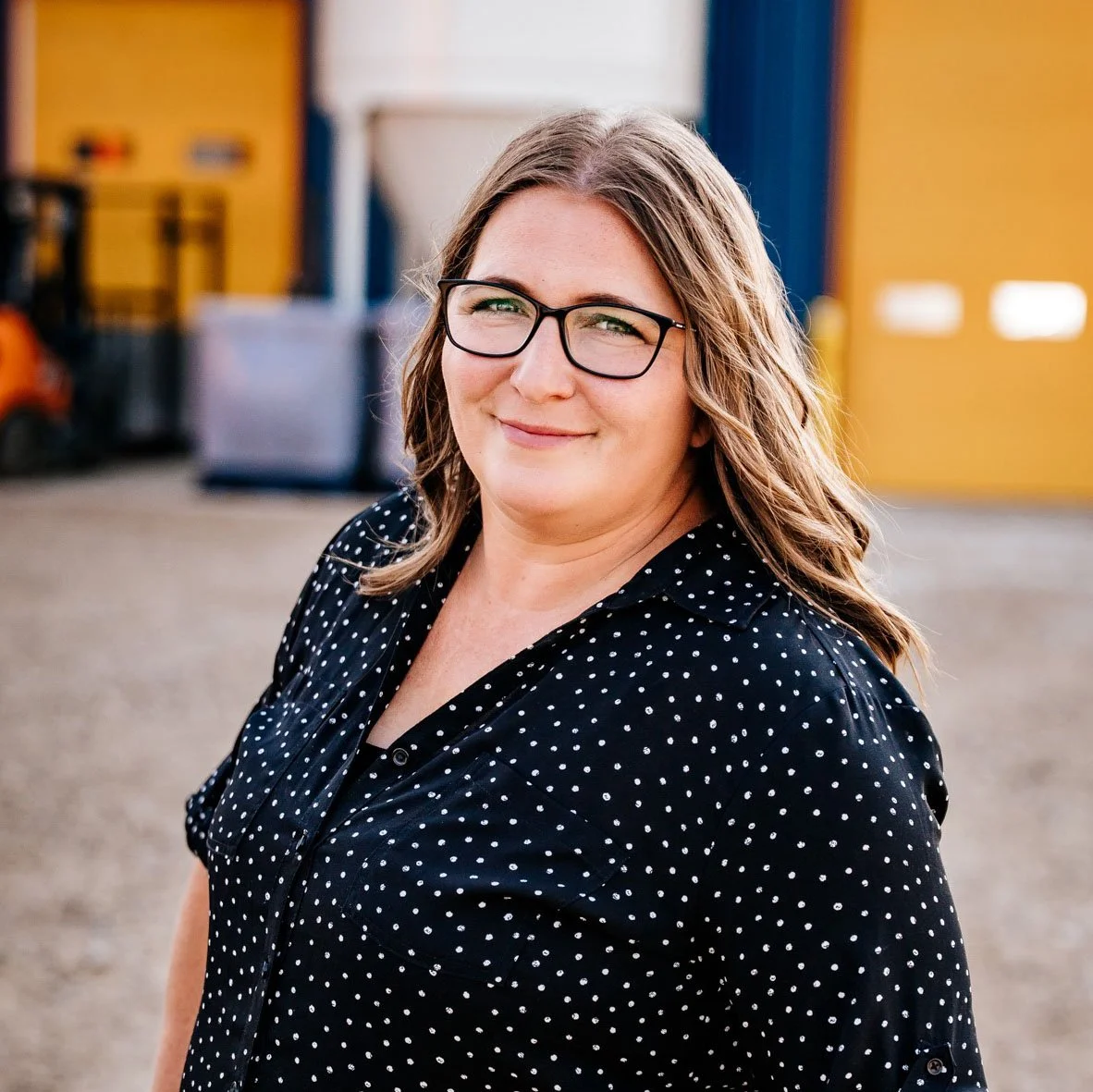 A woman with glasses and wavy hair smiling outdoors in front of a colorful background.