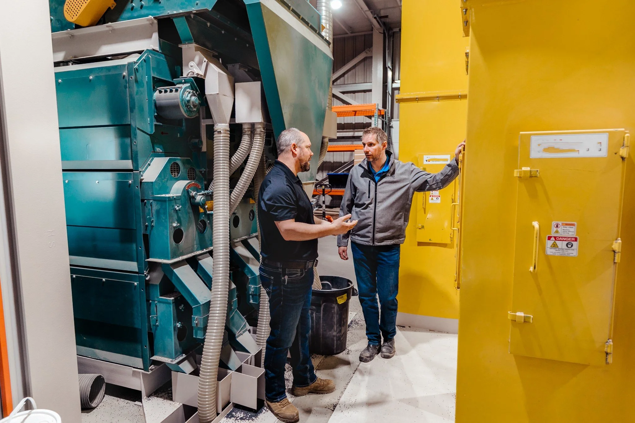 Two men in conversation inside an industrial facility near large blue and yellow machinery, with various technical labels and warning signs.