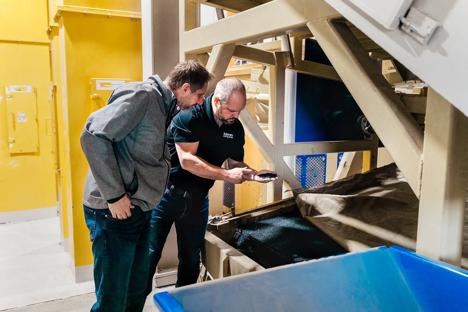 Two men inspecting a piece of machinery in an industrial setting, one holding a black object, likely a tool or part, as they look closely at the equipment.