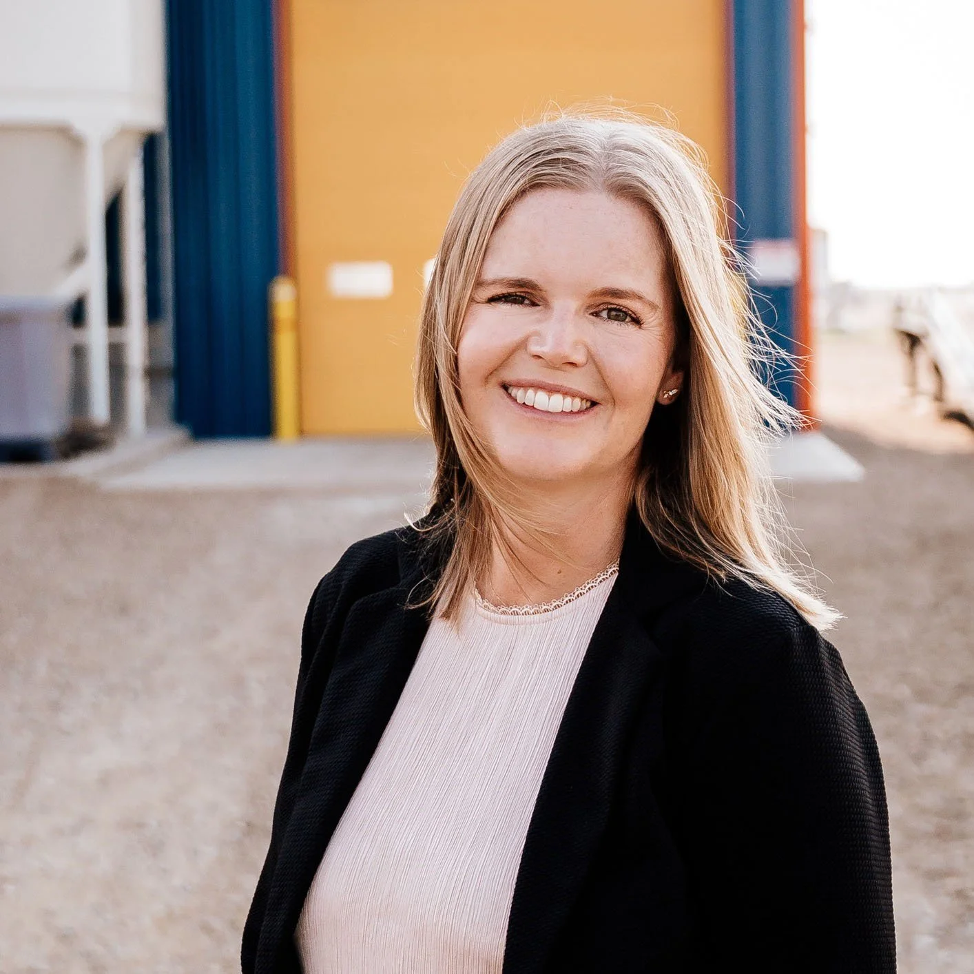 A smiling woman with shoulder-length blond hair wearing a black blazer and a light-colored top, standing outdoors with a colorful building in the background.