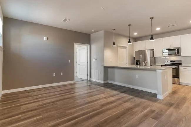 Sala vacía con paredes grises, suelo de madera y cocina moderna con electrodomésticos de acero inoxidable y lámparas colgantes