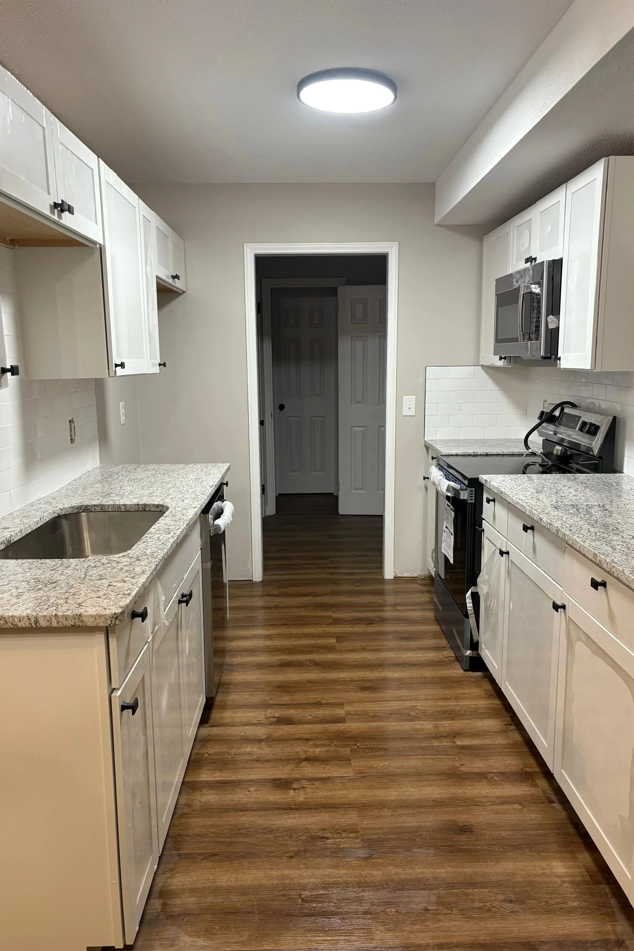 Cocina moderna con gabinetes blancos, encimeras de granito, electrodomésticos de acero inoxidable y suelo de madera.