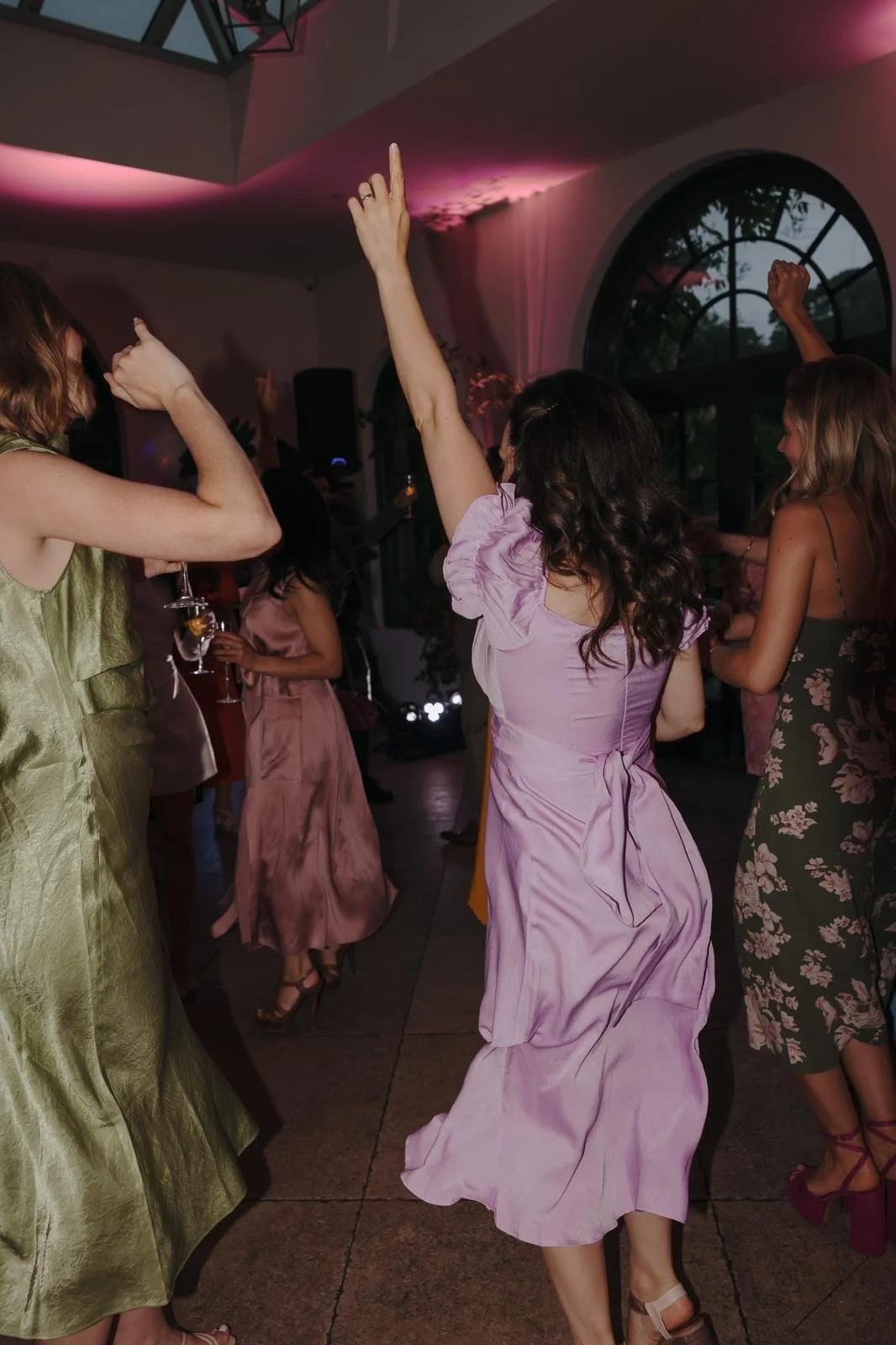 Women dancing at a party or celebration, some raising their hands, inside a decorated venue with pink lighting and large arched windows
